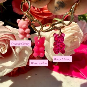 May include: A gold-toned chain bracelet featuring three gummy bear charms in shades of pink. The charms are labelled with text: Creamy Cherry, Watermelon, and Berry Cherry. The bracelet is held up against a background of light pink roses.