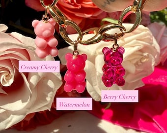 Könnte beinhalten: Ein goldfarbenes Kettenarmband mit drei Gummibärchen-Anhängern in verschiedenen Rosatönen. Die Anhänger sind mit den Texten Creamy Cherry, Watermelon und Berry Cherry beschriftet. Das Armband wird vor hellrosa Rosen gehalten.