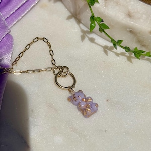 May include: A gold-toned chain necklace featuring a small, light purple gummy bear pendant. The bear-shaped charm is translucent with embedded gold-coloured flakes throughout its surface. The pendant hangs from a circular gold-toned metal ring.