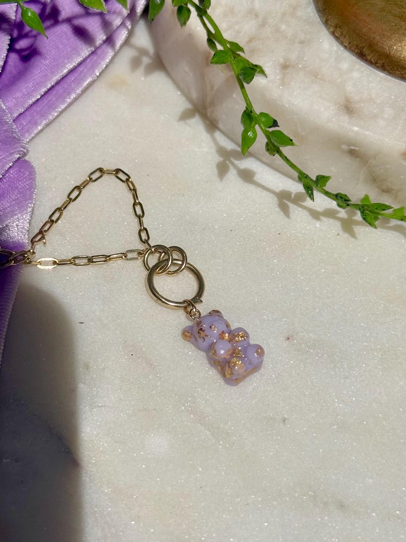 May include: A gold-toned chain necklace featuring a small, light purple gummy bear pendant. The bear-shaped charm is translucent with embedded gold-coloured flakes throughout its surface. The pendant hangs from a circular gold-toned metal ring.