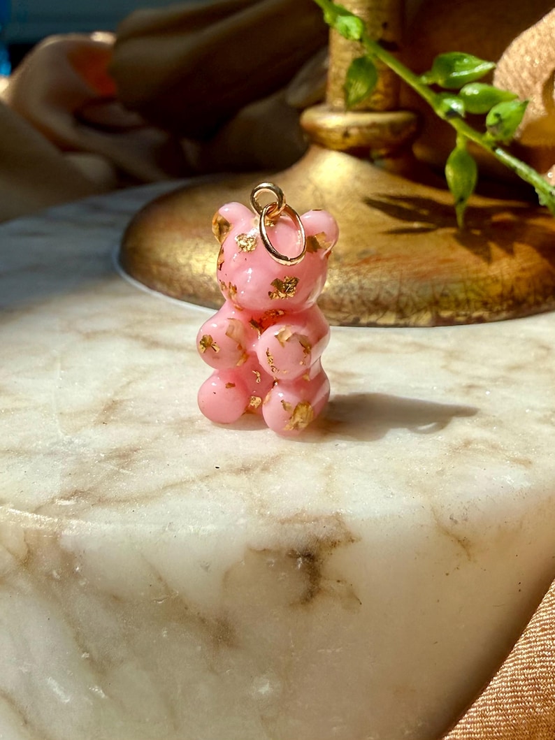 May include: A small, translucent pink gummy bear pendant featuring embedded gold-coloured flakes throughout the resin material. The bear figure has a smooth, glossy finish and includes a circular gold-toned metal jump ring attached to the top of its head.
