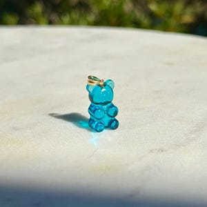 Puede incluir: Un pequeño dije de osito de goma de color turquesa translúcido con una fianza dorada. El dije es una réplica en miniatura de un caramelo de osito de goma, con detalles visibles como los brazos, las piernas y las orejas.
