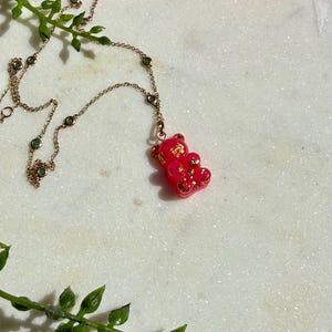May include: A necklace featuring a pink gummy bear pendant with embedded gold flakes, suspended from a delicate gold-tone chain accented with small, round, green stones spaced evenly along the length of the metal link chain.