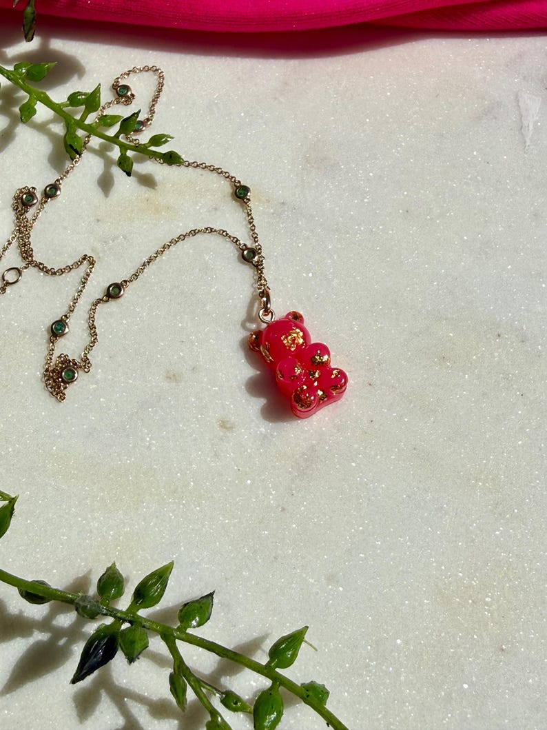 May include: A necklace featuring a pink gummy bear pendant with embedded gold flakes, suspended from a delicate gold-tone chain accented with small, round, green stones spaced evenly along the length of the metal link chain.