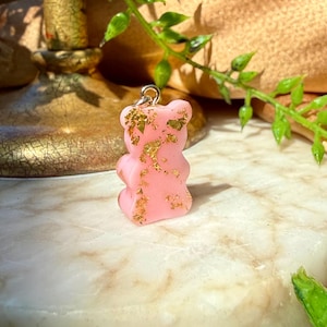 May include: A small, opaque light pink gummy bear-shaped pendant featuring embedded gold-coloured metallic flakes throughout its surface. The pendant includes a silver-toned metal jump ring attached to the top, designed for use as a charm or jewellery component.
