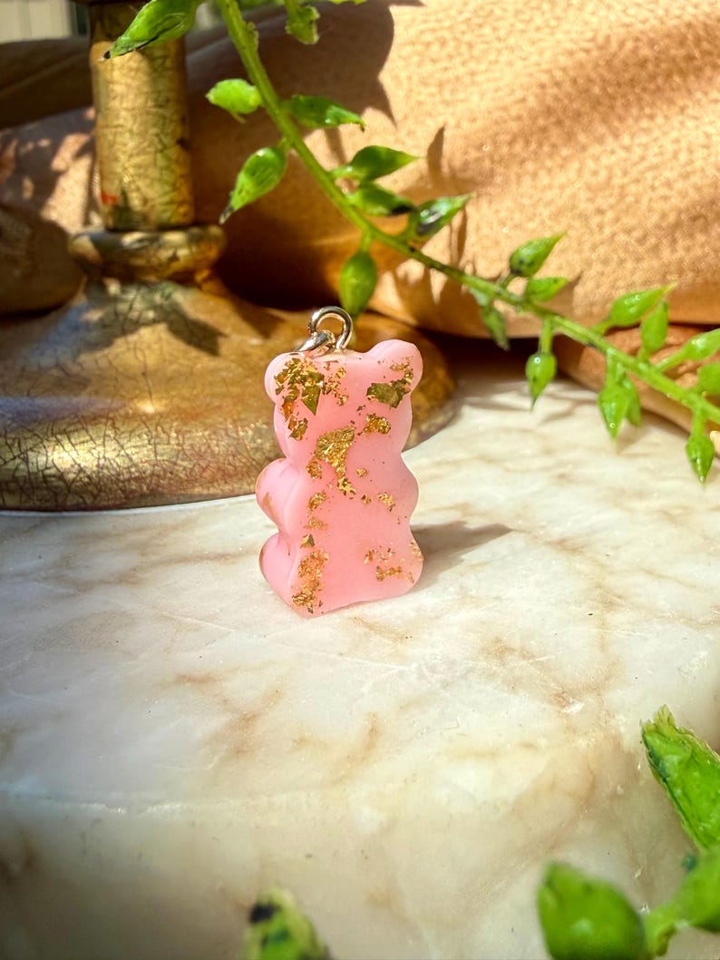 May include: A small, opaque light pink gummy bear-shaped pendant featuring embedded gold-coloured metallic flakes throughout its surface. The pendant includes a silver-toned metal jump ring attached to the top, designed for use as a charm or jewellery component.