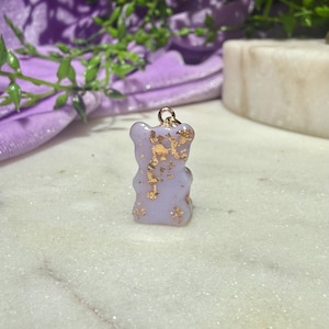 May include: A small, light purple gummy bear-shaped pendant featuring embedded gold leaf flakes throughout the translucent material. The charm includes a small gold-toned metal jump ring attached to the top, designed for use as a jewellery component.