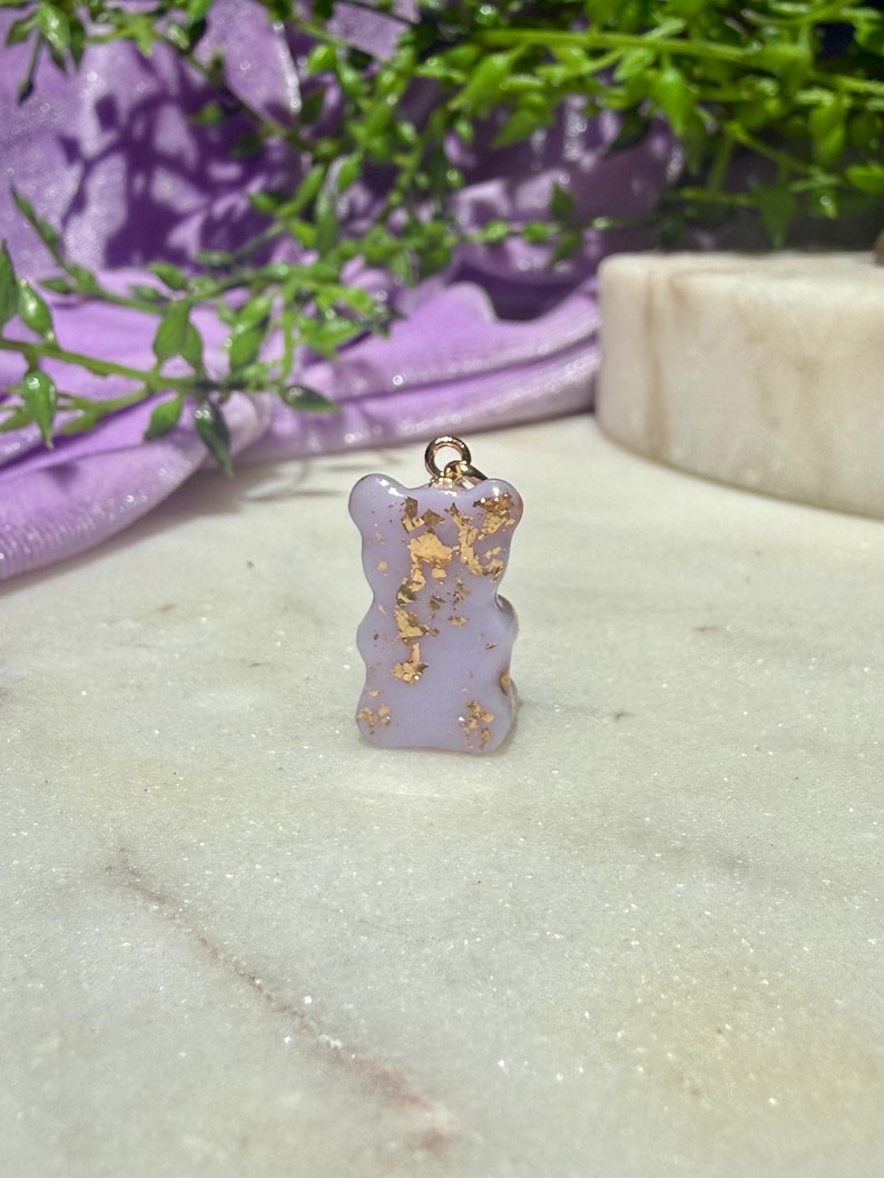 May include: A small, light purple gummy bear-shaped pendant featuring embedded gold leaf flakes throughout the translucent material. The charm includes a small gold-toned metal jump ring attached to the top, designed for use as a jewellery component.