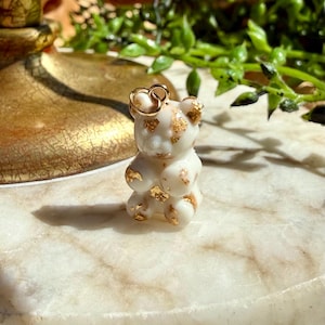 May include: A small, opaque white bear-shaped charm featuring embedded gold-coloured foil flakes throughout its surface. The charm includes a small, circular gold-toned metal jump ring attached to the top of the bear's head for hanging purposes.