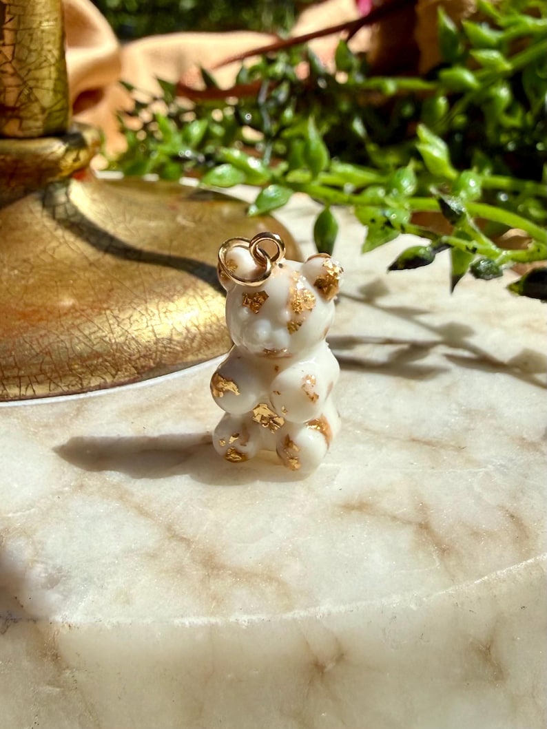 May include: A small, opaque white bear-shaped charm featuring embedded gold-coloured foil flakes throughout its surface. The charm includes a small, circular gold-toned metal jump ring attached to the top of the bear's head for hanging purposes.