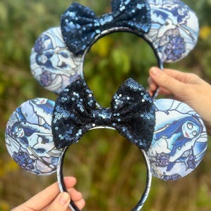 May include: Two headbands featuring round ears with a blue and white printed fabric pattern depicting a female character with long blue hair. Each headband is topped with a centered, dark blue sequined bow. The fabric design includes floral elements.