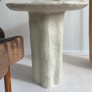 May include: A textured, off-white side table with a unique, organic shape. The table has a round top and a pedestal base. The table is approximately 24 inches tall and 20 inches wide.