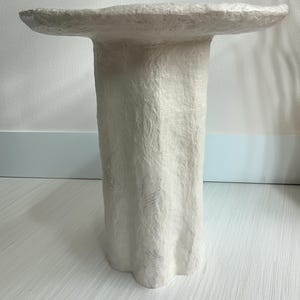 Puede incluir: Una mesa pedestal blanca y texturizada con una parte superior redonda y una base acanalada. La mesa mide aproximadamente 76 cm de altura y está hecha de un material de color claro, posiblemente papel o yeso.