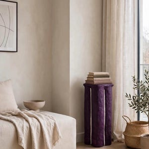 May include: A tall, cylindrical pedestal with a deep purple, textured finish and vertical white recessed channels. The top surface of the pedestal supports a stack of three books with neutral-colored covers, positioned in a corner near a window.