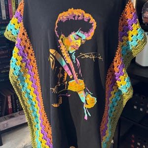 May include: Black t-shirt featuring a colorful illustration of Jimi Hendrix playing guitar, with his signature. The sleeves are adorned with a crocheted granny square design in shades of green, orange, purple, and yellow.