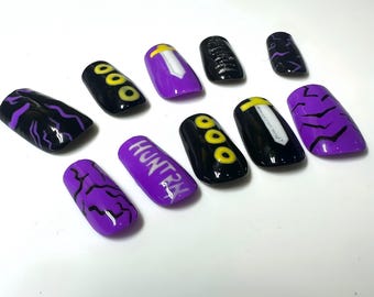 May include: A collection of ten artificial nails in various designs. The nails feature a glossy finish with designs in black, purple, and yellow. Some nails have wavy patterns, while others have text or geometric shapes. The nails are arranged on a white surface.