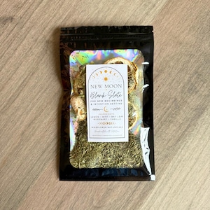 May include: A black resealable pouch with a holographic window containing dried herbs and citrus slices. A white label features the text NEW MOON Blank Slate FOR NEW-BEGINNINGS & INTENTION SETTING, listing ingredients: LEMON, MINT, BAY LEAF, ROSEMARY, VANILLA.
