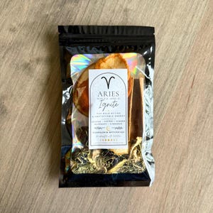 Puede incluir: Una bolsa negra resellable que contiene una mezcla de botánicos. La bolsa presenta una etiqueta con la palabra "ARIES" y la palabra "Ignite". Los ingredientes enumerados son naranja, cereza, jengibre, romero y canela. La bolsa contiene rodajas de naranja secas y palitos de canela.