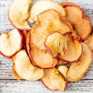 Może przedstawiać: Zbliżenie na suszone plasterki jabłka, w jasnobrązowym kolorze z czerwonawymi krawędziami. Owoce są ułożone na białej drewnianej powierzchni. Obraz skupia się na fakturze i kolorze suszonych jabłek, dając naturalny i rustykalny wygląd.