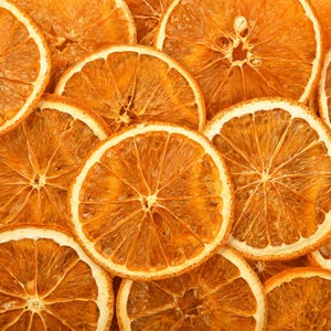 Könnte beinhalten: Nahaufnahme von getrockneten Orangenscheiben, die einen leuchtenden Orangeton mit einem helleren, fast weißen Außenring aufweisen. Die Scheiben sind in einem geschichteten Muster angeordnet, das ihre kreisförmige Form und strukturierten Oberflächen hervorhebt.