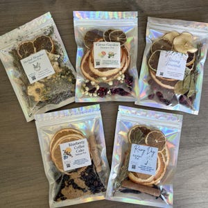 Op de afbeelding: Vijf transparante zakjes gevuld met gedroogde planten en schijfjes citrusvruchten. Elk zakje heeft een label met een andere naam: "Herbal Meadow", "Citrus Garden", "Spring Forward", "Blueberry Coffee Cake" en "Rainy Day". De zakjes liggen op een houten oppervlak.