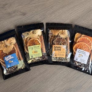 May include: Four black resealable pouches containing dried fruit, cinnamon sticks, and spices. Labels read Sweeter Weather, Cozy Harvest, Spooky Season, and Pumpkin Spice Latte. Each pouch displays a unique blend of dehydrated orange slices, apple pieces, and aromatic botanicals.