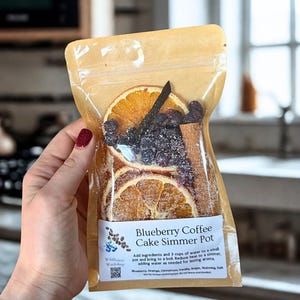 May include: A clear bag containing ingredients for a Blueberry Coffee Cake Simmer Pot. The bag includes dried orange slices, blueberries, a cinnamon stick, and a vanilla bean. The label reads "Blueberry Coffee Cake Simmer Pot".