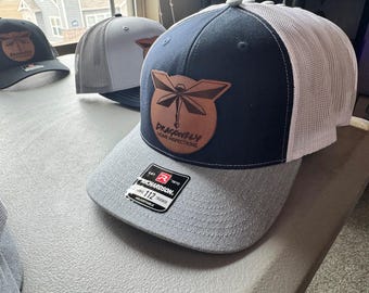 May include: A navy blue and gray trucker hat with a white mesh back. The hat has a brown leather patch with a dragonfly design and the text "Dragonfly Home Inspections".
