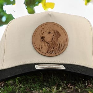 May include: A white baseball cap with a black brim. A round, brown leather patch with a dog's face engraved on it is attached to the front of the cap. The dog's name, "LOU", is engraved below the image.