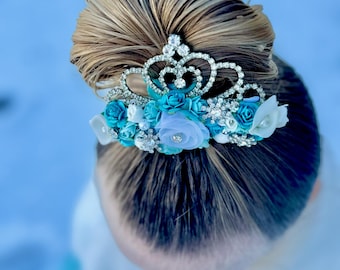 May include: A silver tiara with rhinestones and blue and white flowers, worn in a bun of blonde hair.