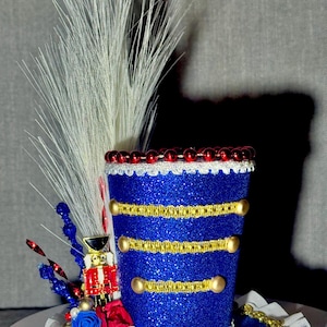 Puede incluir: Un sombrero de copa decorativo azul brillante con adornos y detalles dorados. El sombrero presenta un soldado de juguete en miniatura, adornos florales rojos y azules y detalles de plumas blancas. El sombrero es probablemente para una fiesta.