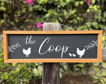 May include: A rectangular wooden sign with a black background and the words "the Coop" in white script. The sign features white chicken silhouettes and floral accents, framed by a light brown wooden border. The sign is mounted on a wooden post.