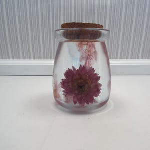 Op de afbeelding: Een heldere glazen pot met een kurken stopper, met daarin een gedroogde, dieproze bloem. De bloem staat in het midden van de pot, omringd door een heldere vloeistof. De pot staat op een wit oppervlak.