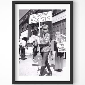 May include: Black and white framed photograph of a protest. A woman holds a sign that reads "SCREW SEXISTS." Other protesters hold signs with text. The image is a historical document of a social movement.