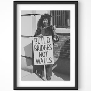 May include: Monochrome framed print showing a person holding a sign with the text "BUILD BRIDGES NOT WALLS." The person is standing outside a building. The image is a portrait.
