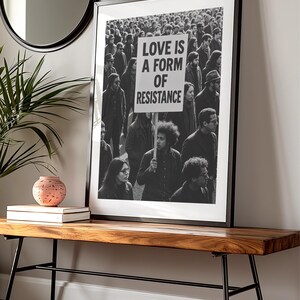 Pu&ograve; includere: Stampa in bianco e nero incorniciata con una folla di persone. Un cartello nella folla recita "LOVE IS A FORM OF RESISTANCE". La stampa &egrave; esposta su una panca di legno con gambe in metallo nero, con uno specchio rotondo e oggetti decorativi sullo sfondo.