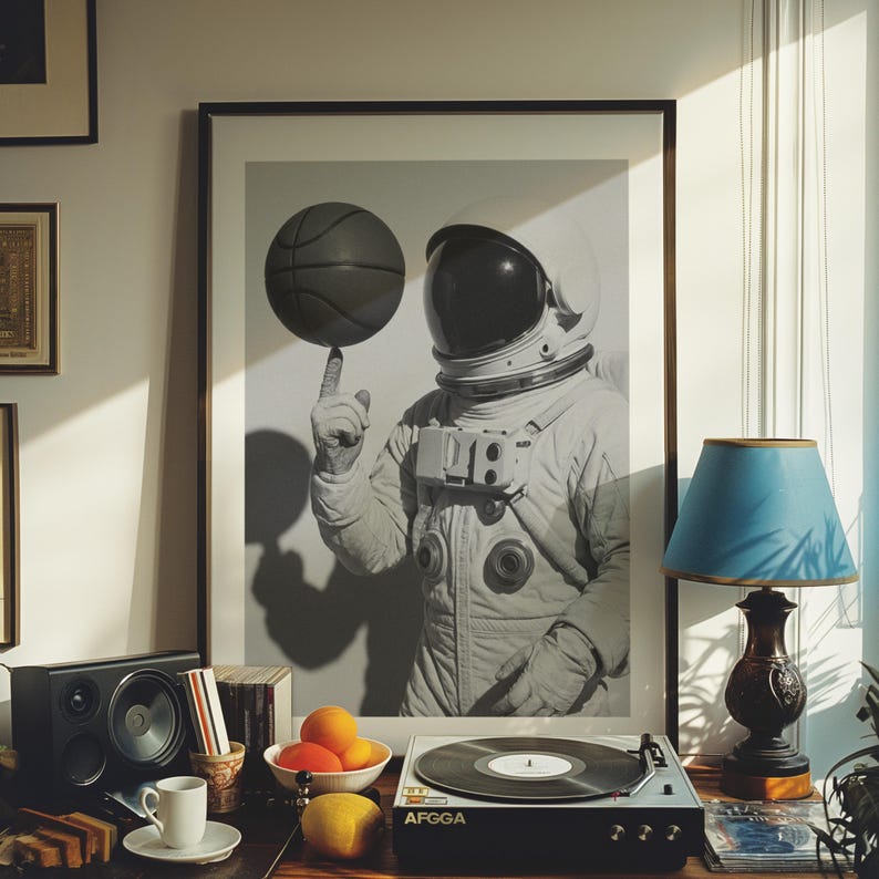 May include: A framed black and white print of an astronaut balancing a basketball on a finger. The print is displayed on a wooden table with a record player, speakers, fruit, and a lamp with a blue lampshade. A cup and saucer are also on the table.