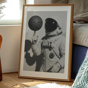 May include: Black and white framed print of an astronaut balancing a basketball on a finger. The astronaut is wearing a white spacesuit and helmet. The print is displayed in a wooden frame, leaning against a wall.