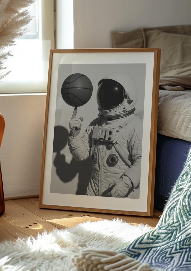 May include: Black and white framed print of an astronaut balancing a basketball on a finger. The astronaut is wearing a white spacesuit and helmet. The print is displayed in a wooden frame, leaning against a wall.