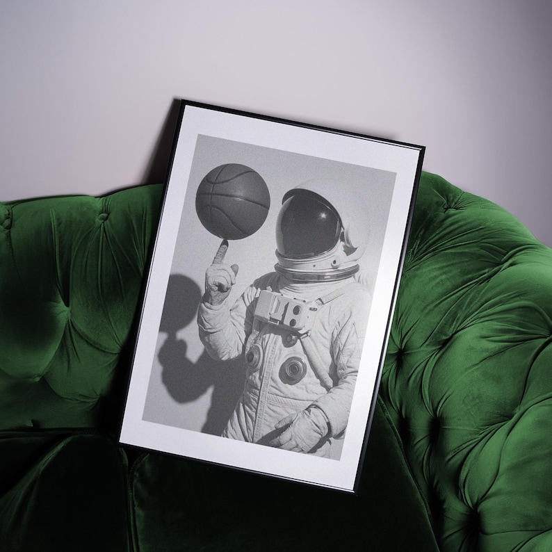 May include: Black and white framed print featuring an astronaut balancing a basketball on their finger. The astronaut is wearing a white spacesuit and helmet. The image is displayed on a green velvet couch.