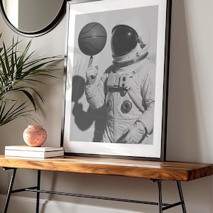 May include: Black and white framed print of an astronaut balancing a basketball on a finger. The astronaut is wearing a spacesuit and helmet. The print is displayed on a wooden bench with black metal legs.