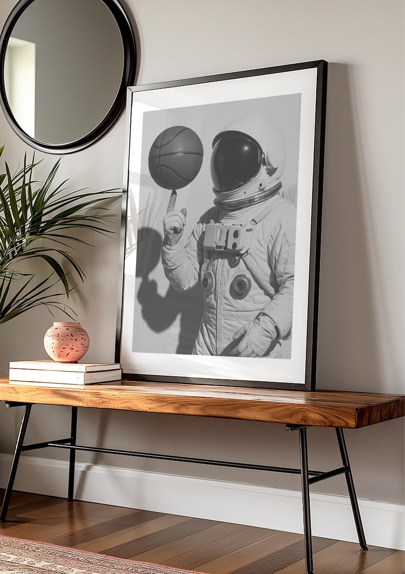 May include: Black and white framed print of an astronaut balancing a basketball on a finger. The astronaut is wearing a spacesuit and helmet. The print is displayed on a wooden bench with black metal legs.