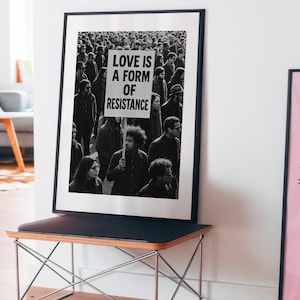 Pu&ograve; includere: Stampa in bianco e nero incorniciata con una folla. Un cartello nella folla recita "LOVE IS A FORM OF RESISTANCE." La stampa &egrave; esposta su una panca nera con una cornice in metallo.
