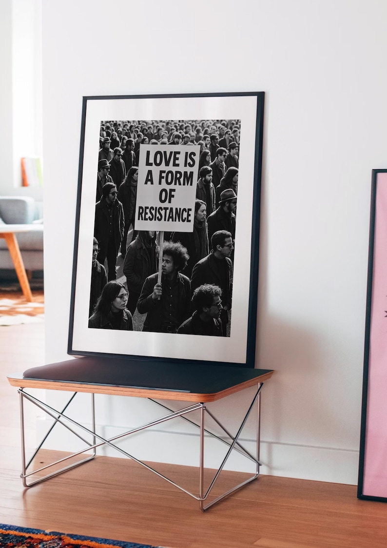 Pu&ograve; includere: Stampa in bianco e nero incorniciata con una folla. Un cartello nella folla recita "LOVE IS A FORM OF RESISTANCE." La stampa &egrave; esposta su una panca nera con una cornice in metallo.