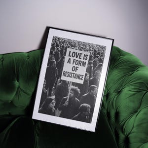 Pu&ograve; includere: Stampa in bianco e nero incorniciata con una folla di persone. Un cartello nella folla recita "LOVE IS A FORM OF RESISTANCE". La stampa &egrave; esposta su un divano di velluto verde scuro.