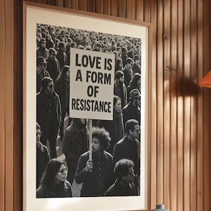 Pu&ograve; includere: Stampa incorniciata in bianco e nero con una folla di persone, con un cartello con la scritta "LOVE IS A FORM OF RESISTANCE". La stampa &egrave; esposta su una parete di legno, con un vaso decorativo.