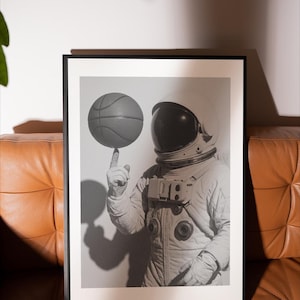 May include: Black and white framed print of an astronaut balancing a basketball on their fingertip. The astronaut is wearing a spacesuit and helmet. The image is displayed on a white background, with a brown leather couch in the foreground.