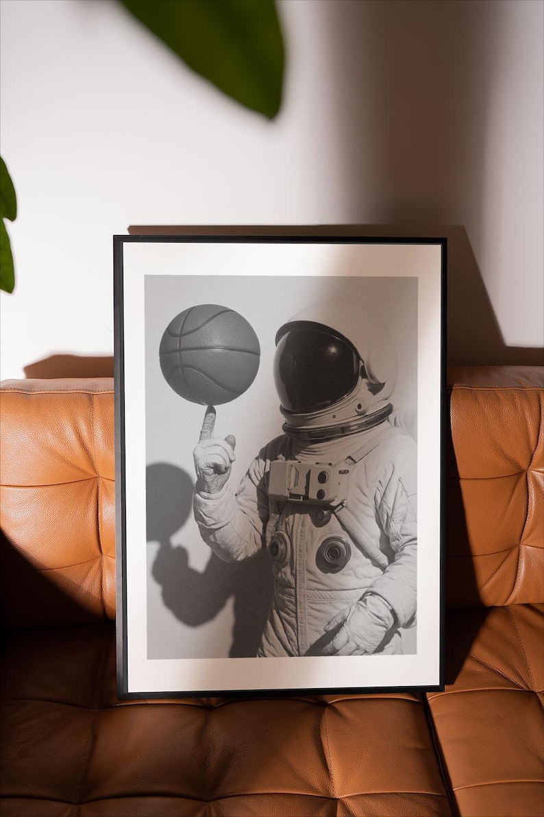 May include: Black and white framed print of an astronaut balancing a basketball on their fingertip. The astronaut is wearing a spacesuit and helmet. The image is displayed on a white background, with a brown leather couch in the foreground.