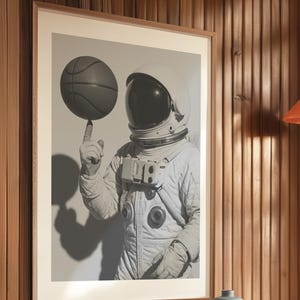 May include: Framed black and white print of an astronaut balancing a basketball on their finger. The astronaut is wearing a white spacesuit and helmet. The image is set against a neutral background.