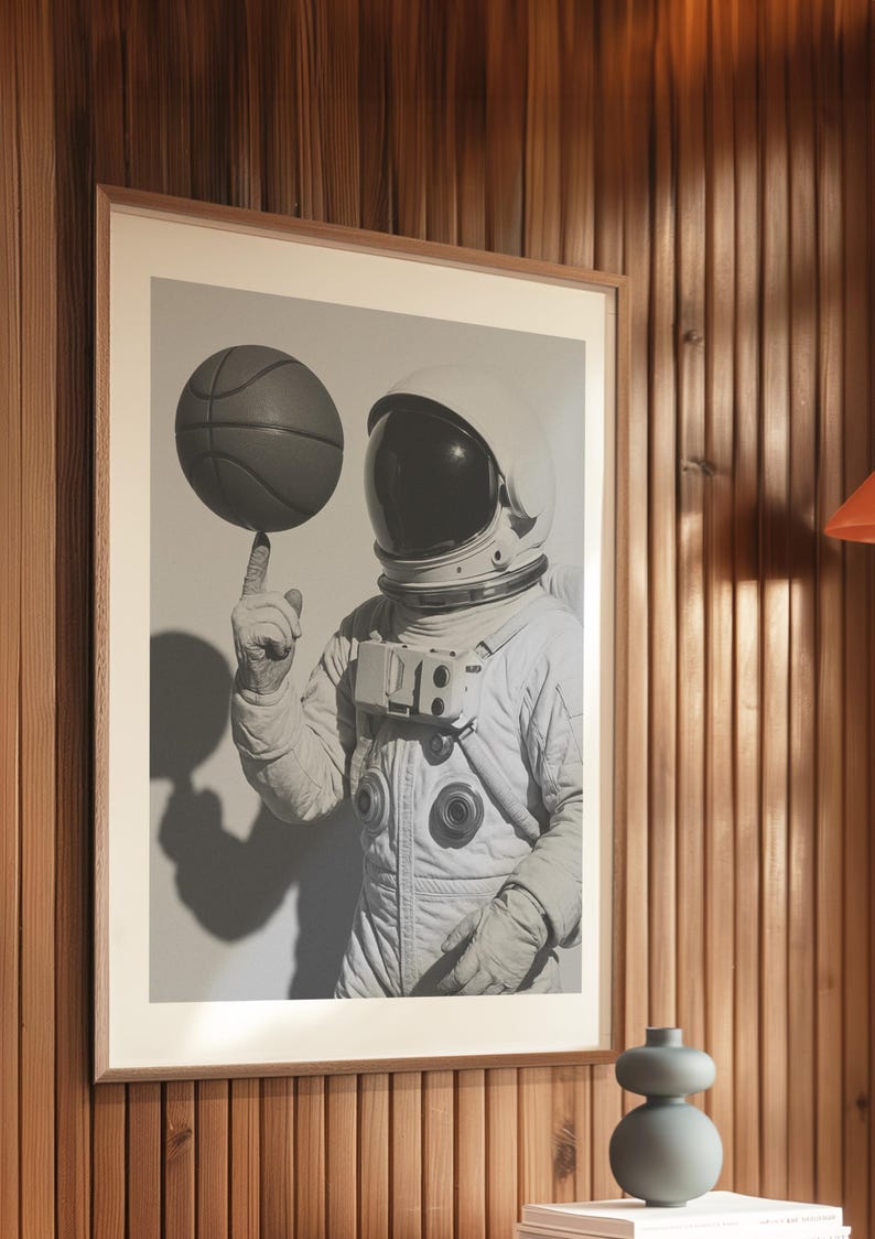 May include: Framed black and white print of an astronaut balancing a basketball on their finger. The astronaut is wearing a white spacesuit and helmet. The image is set against a neutral background.